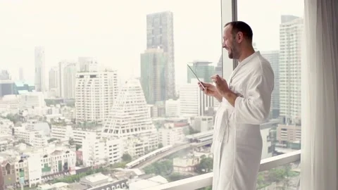 Happy man in bathrobe browsing tablet standing by window, super slow motion Stock Footage 127973375