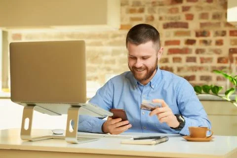 A happy man with a beard using a smartphone in front of the computer at home. Stock Photos