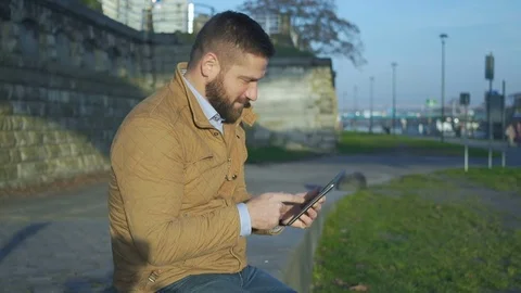 Happy man browsing tablet computer, sitting on wall at boulevard next to river Vídeo Stock 72775421