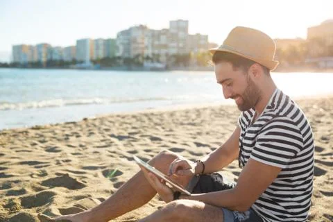 Happy man in hat sitting on beach using tablet Foto stock
