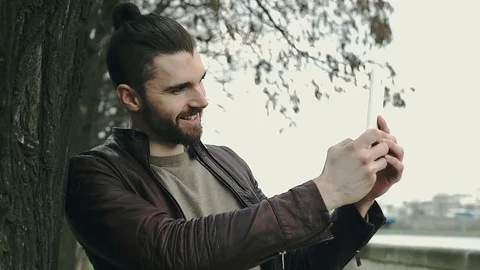 Happy man in leather jacket leaning on the tree and doing selfies, steadycam sho Stock Footage