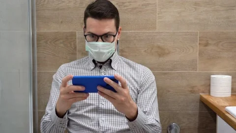 Happy man in protective face mask sit in toilet, hold phone, plays in game Stock Footage 131463314
