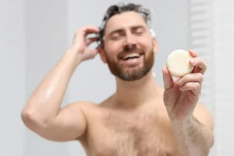 Happy man showing solid shampoo bar in bathroom, selective focus Stock Photos