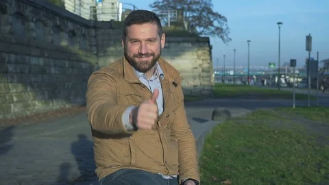 Happy man shows ok at camera, sitting on wall at boulevard next to river Vidéo 72775160