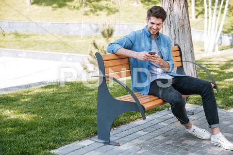 Happy Man Sitting On The Bench Outdoors And Using Smartphone