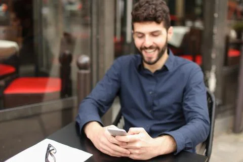 Happy man sitting at cafe table and chatting by smartphone with Stock Photos