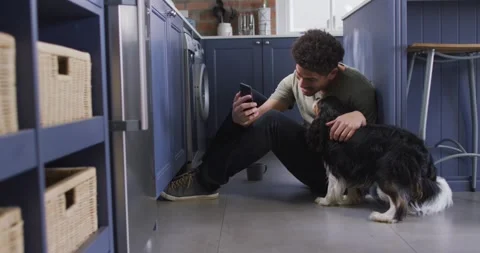 Happy man sitting on floor in kitchen with his pet dog, making video call using Stock Footage 340985754