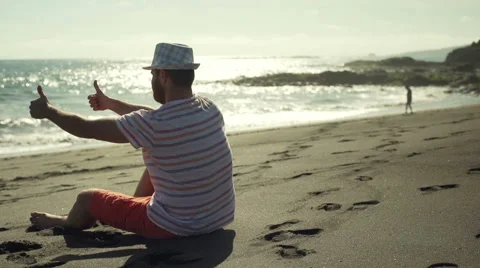 Happy man sitting on the sandy beach and showing thumbs, steadycam shot 스톡 동영상 60132675