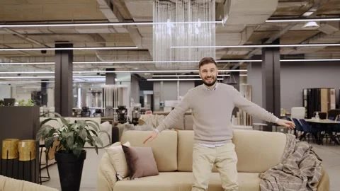 Happy man standing behind a sofa in furniture store, showing new interior design Stock Footage 334654222