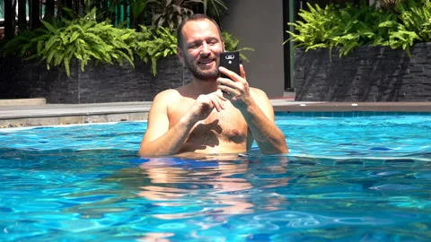 Happy man standing in the pool and having a videocall on smartphone, slow motion Stock Footage 88538553