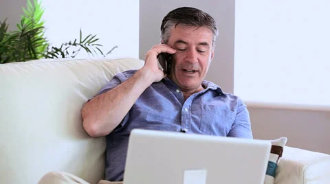 Happy man talking on the phone on the sofa with his laptop Stock Footage