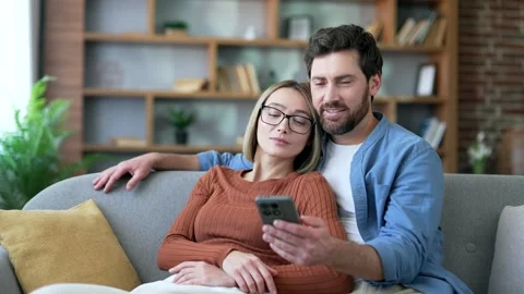 Happy married couple using mobile phone resting sitting on sofa in living room  Stock Footage 337929949