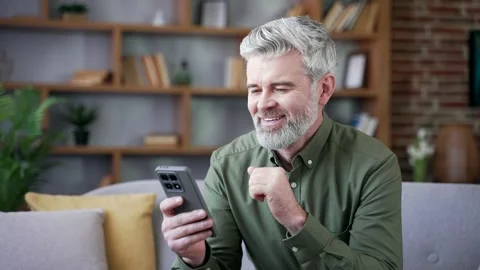 Happy mature bearded man with gray hair sitting on sofa at home using phone.  Stock Footage 328706653