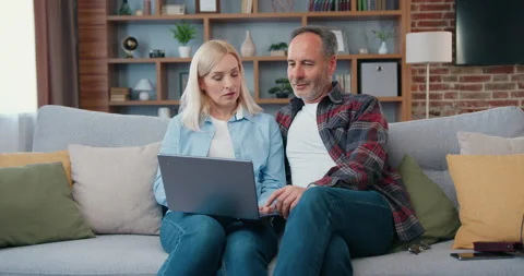 Happy middle aged couple looking at laptop screen sitting on sofa at home. Woman Stock Footage 194137971