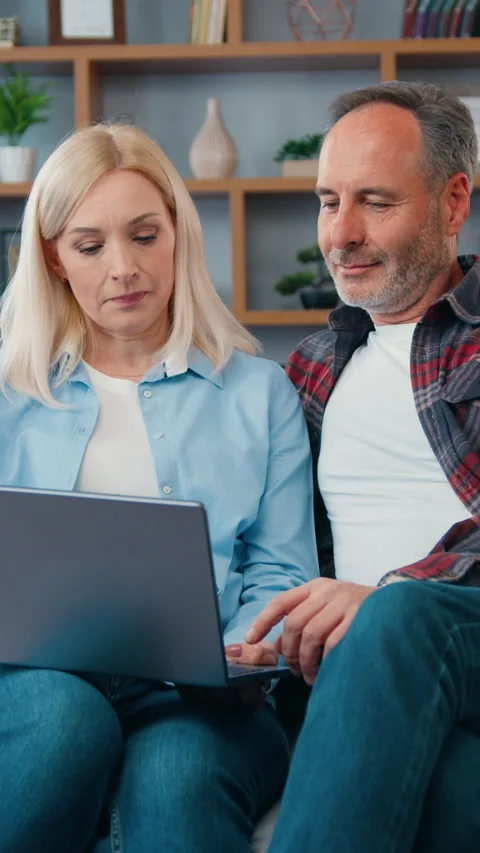 Happy middle aged couple looking at laptop screen sitting on sofa at home. Woman Stock Footage 334935809