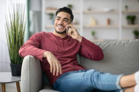 Happy middle-eastern guy reclining on couch, talking on phone Stock Photos