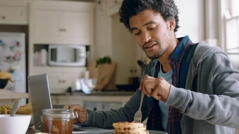 Happy mixed race man using laptop computer working in kitchen eating waffles for Stock Footage 109511303