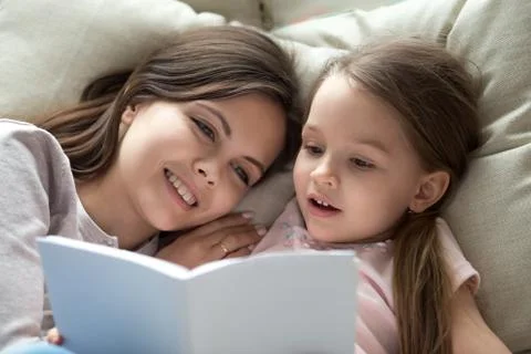 Happy mom listening child daughter learning reading book in bed Stock Photos