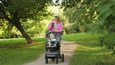 Happy mother drinks coffee while walking in the summer park with her young Stock Footage 234314510