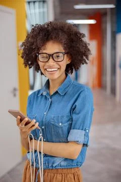 Happy multiethnic woman posing with mobile phone in the office corridor Stock Photos