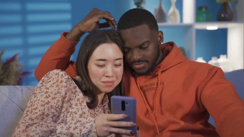 Happy multiracial couple sitting on sofa at home chatting, looking at phone Stock Footage 228809466