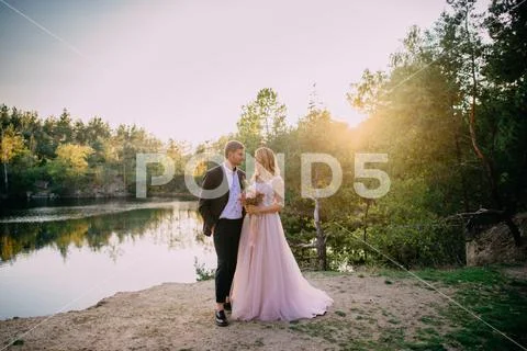 Happy newlyweds standing on the lake shore at sunset Stock Photos