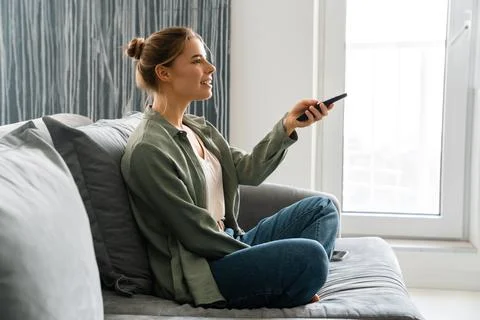 Happy nice woman watching TV with remote control while sitting on sofa Stock Photos