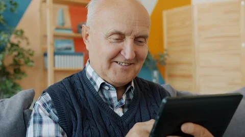 Happy old man enjoying tablet device touching screen smiling relaxing on sofa in Stock Footage 128469017