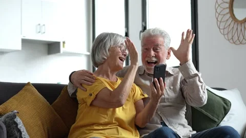 Happy older couple sit on couch staring at cellphone screen gesturing looking ov Stock Footage 237939451