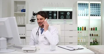 Happy Physician Man Work Talk on Mobile Phone and Use Computer in Hospital Room Stock Footage 85570453