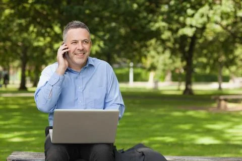 Happy professor talking on phone while sitting on bench using laptop on campus Photos