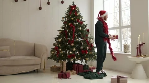 A happy red-bearded young man in a Santa hat unpacking Christmas presents. Stock Footage 294038968
