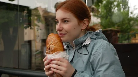 Happy red-haired girl eats a loaf at the bus stop Stock Footage 112973461