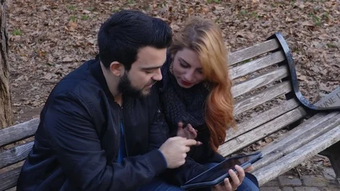 Happy romantic couple with computer tablet, relaxing on bench at the park Stock Footage 74720820