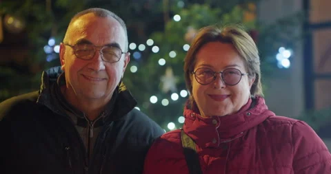 Happy senior couple smiling on camera outside with christmas tree and lights in Stock Footage 286356420