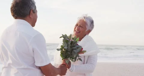 Happy senior hispanic man proposing to senior woman with roses on beach at Stock Footage 341936268