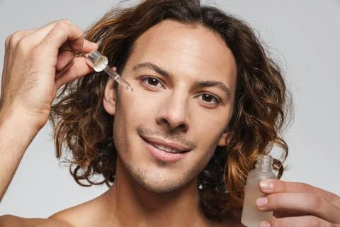 Happy shirtless guy smiling while applying face serum Stock Photos