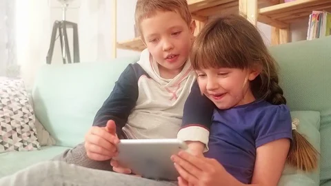 Happy siblings lying on sofa at home and playing with pc tablet together Stock Footage 73511644