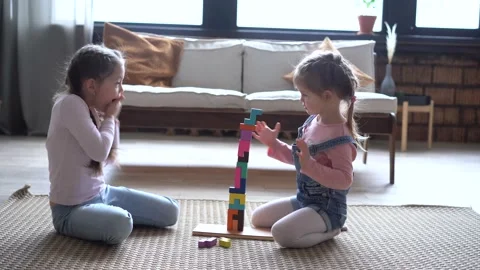Happy sisters sitting on carpet, playing with colorful wooden blocks constructor 库存影片 147388241