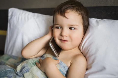 Happy small boy talking mobile phone lying on the bed in morning Stock Photos