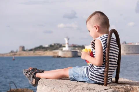 Happy small kid sitting on breakwater on background of sea and lighthouse a.. 스톡 사진