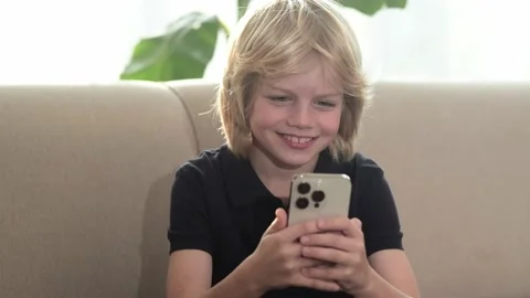 Happy smiling blonde boy using looking phone and sitting home on sofa, holding Stock-Footage 231297501
