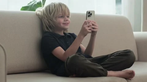 Happy smiling blonde boy using looking phone and sitting home on sofa, holding Stock-Footage 231578435