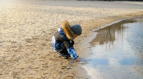 Happy smiling boy touching cold water in pond at park on windy winter day Foto stock