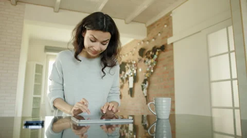 Happy, smiling brunette typing on tablet sitting at table in living room Stock Footage 244324736