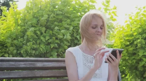 Happy smiling girl using a smart phone in a city park sitting on a bench Stock Footage 64345427