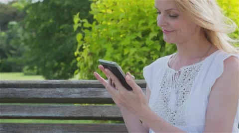 Happy smiling girl using a smart phone in a city park sitting on a bench  Stock Footage 64347794