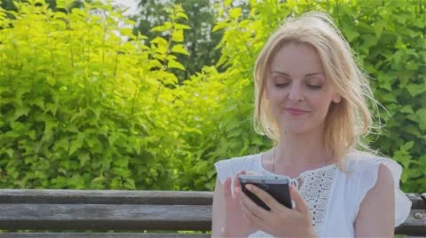 Happy smiling girl using a smart phone in a city park sitting on a bench  Stock Footage 64348707
