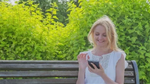 Happy smiling girl using a smart phone in a city park sitting on a bench Stock Footage 64350126