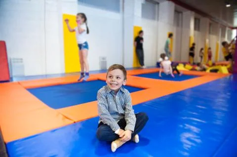 Happy smiling little boy sitting on trampoline among jumping kids indoors Stock-Fotos